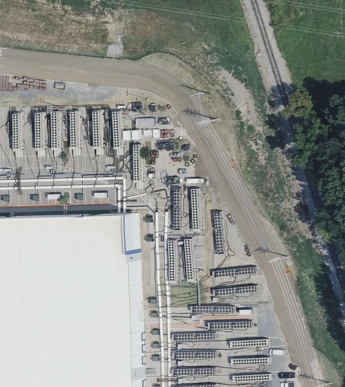 Aerial photo of one corner of the Colossus 2 site. Dozens of long rectangular units, each topped with two rows of round fans, stand in rows beside a large white building, next to a road and a rail line.